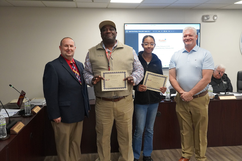 A community member and a student pose with district leaders while holding certificates of recognition in the Bladen County Schools boardroom.