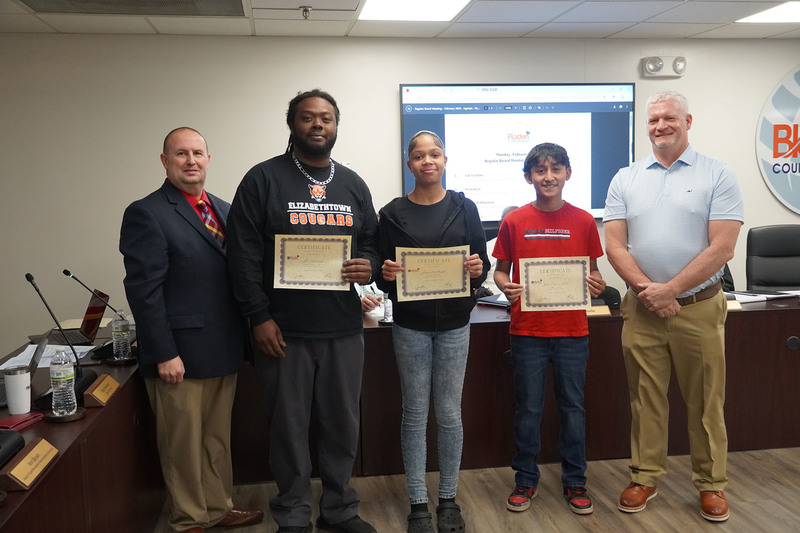 Three students and a school representative stand with district leaders, holding certificates of recognition at a Bladen County Schools Board of Education meeting.