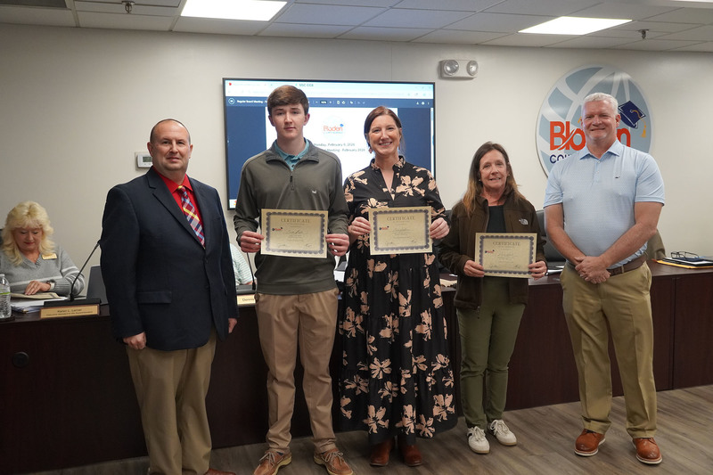 Three individuals, including a student, pose with district leaders while holding certificates of recognition in the Bladen County Schools boardroom.