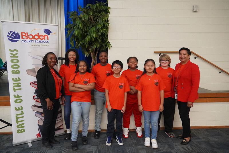 A group of elementary students wearing matching red THS shirts pose with two staff members near a Bladen County Schools “Battle of the Books” banner. The students stand in front of a plant and stage area inside a school auditorium.
