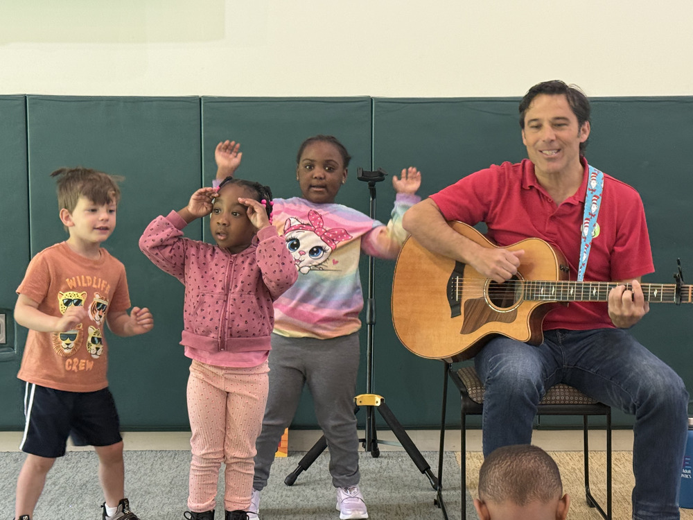 Man playing guitar with three children next ot him singing with hand motions.