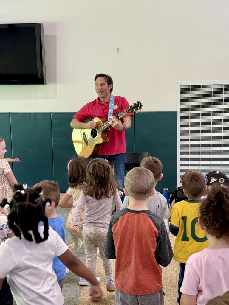 Man playing guitar and singing to group of children standing in front of him.