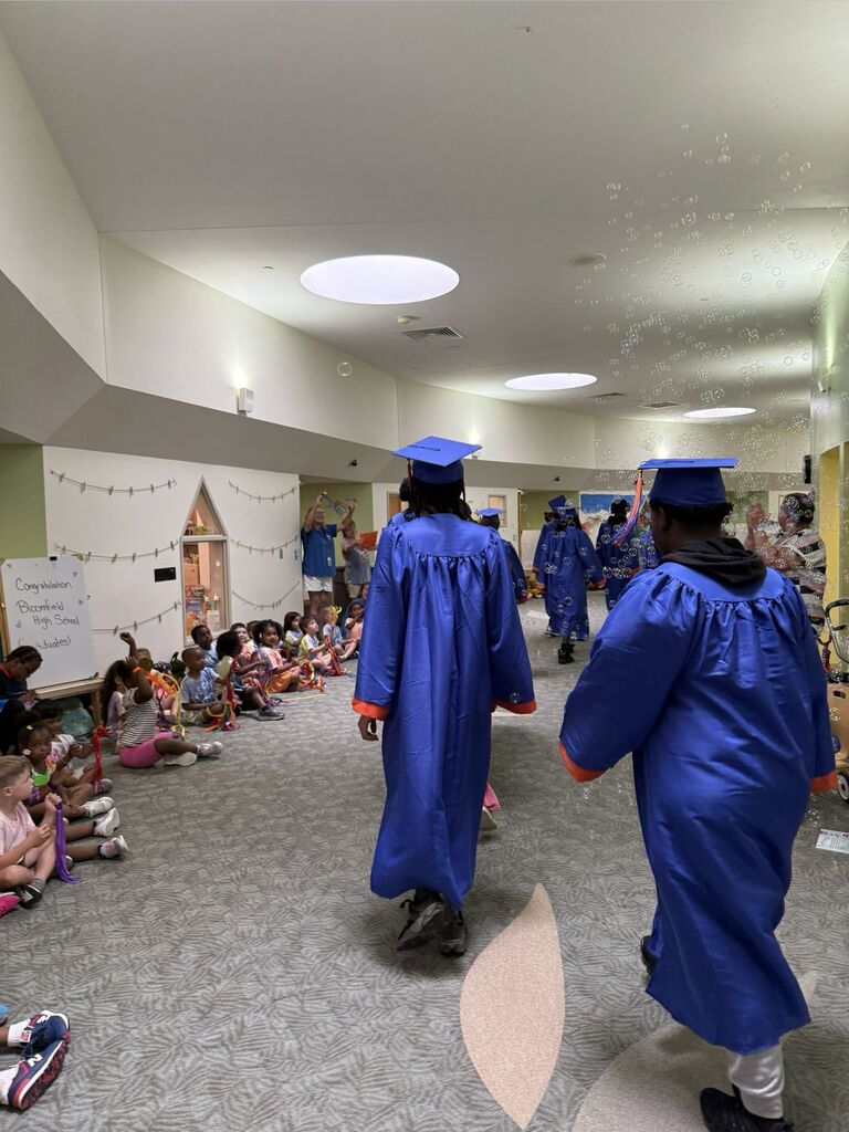 High school students in caps and gowns for graduation walking in hallway with young children cheering for them.