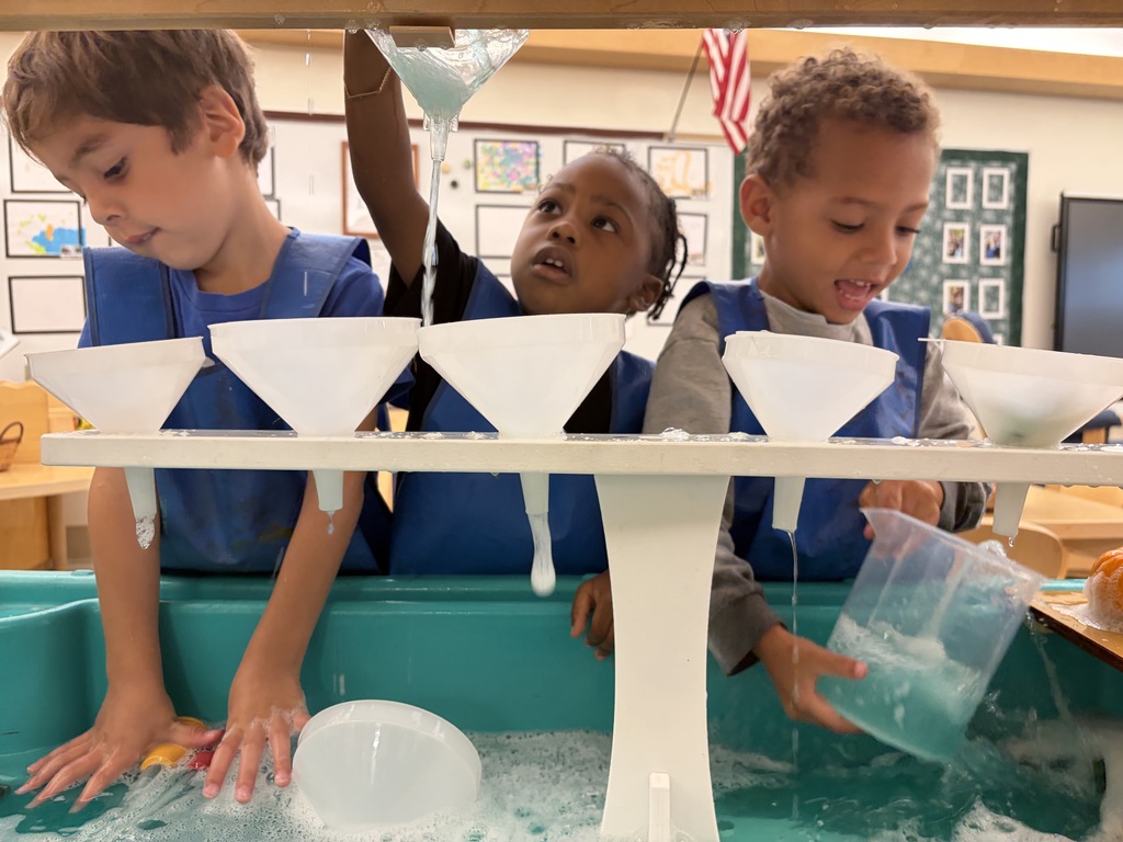 Three preschool boys use funnels and containers to pour water at the water table.