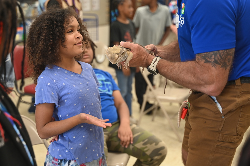 Person holds animal as girl with curly hair and blue shirt stands nearby. Others sit or stand behind.
