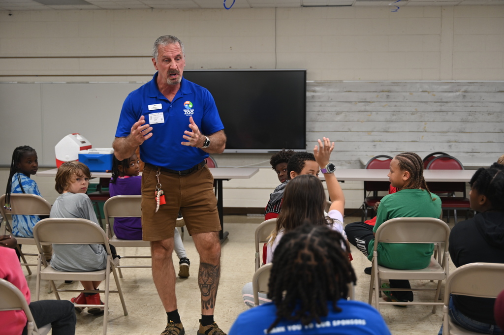 A man in a blue shirt and brown shorts stands in front of seated students in a classroom.