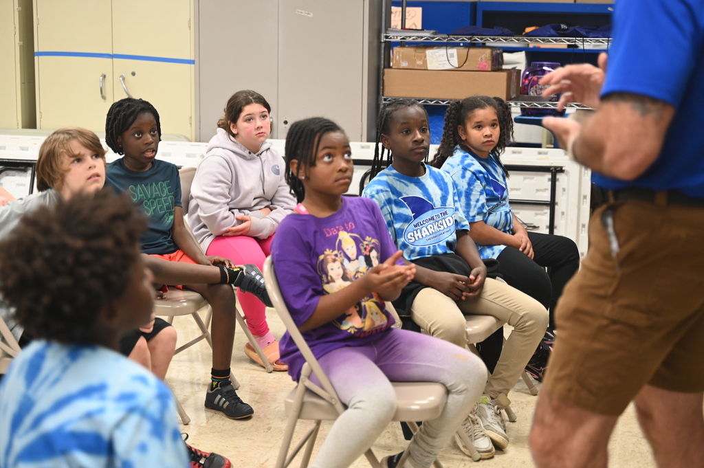 Group of students in chairs, one child clapping, with a man standing in front, explaining with hand gestures.
