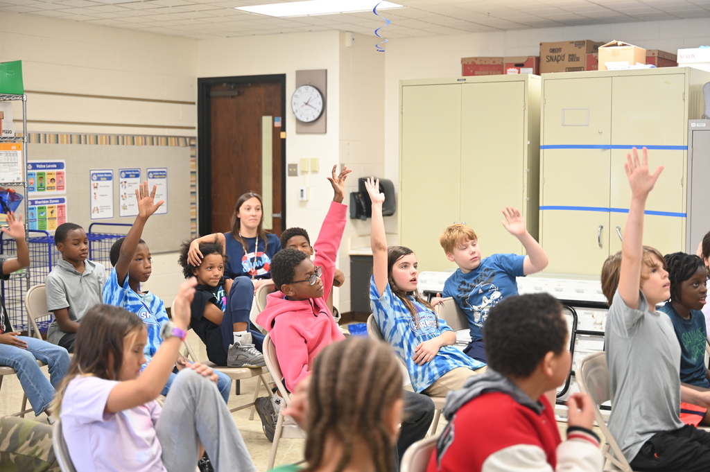 Students in a classroom with arms raised and others seated, near a bulletin board, clock, and storage cabinets.