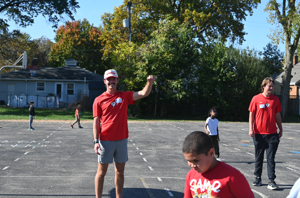 Adults and children in red shirts and shorts in a parking lot. Trees and houses in the background.