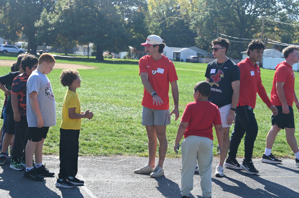 Adults and children in red shirts stand in a circle on a field with a grassy background.