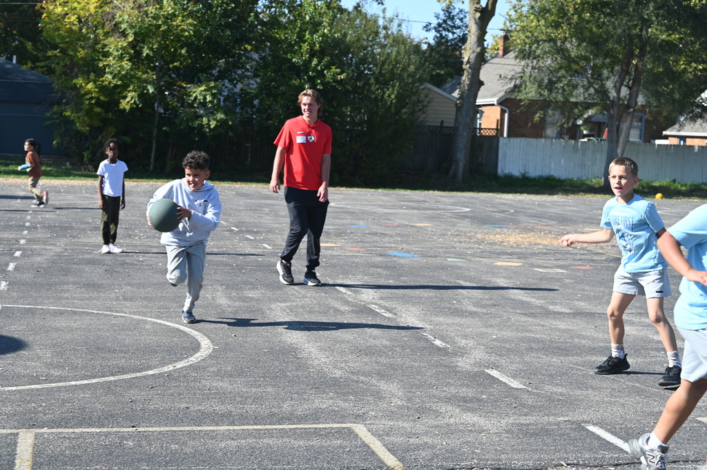 A group of children and adults playing a game outdoors. Some children are running and holding a ball.