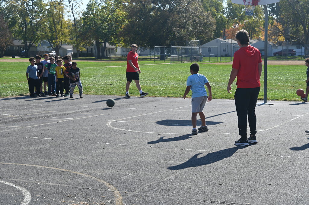 People play kickball on a basketball court. A person in red stands behind a child in white.