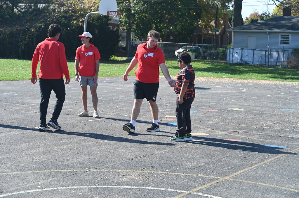 Four adults and a child on a basketball court. Four people in red shirts and black pants, one holding the hand of a child.