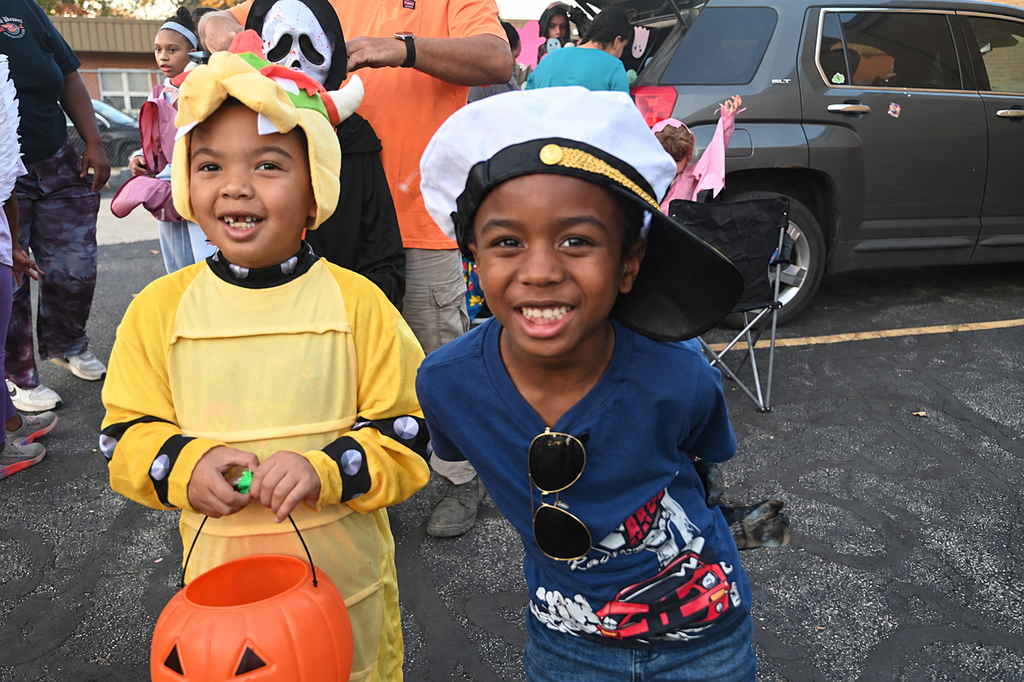 Two young children smiling, one in costume holding a pumpkin, other with hat and sunglasses.
