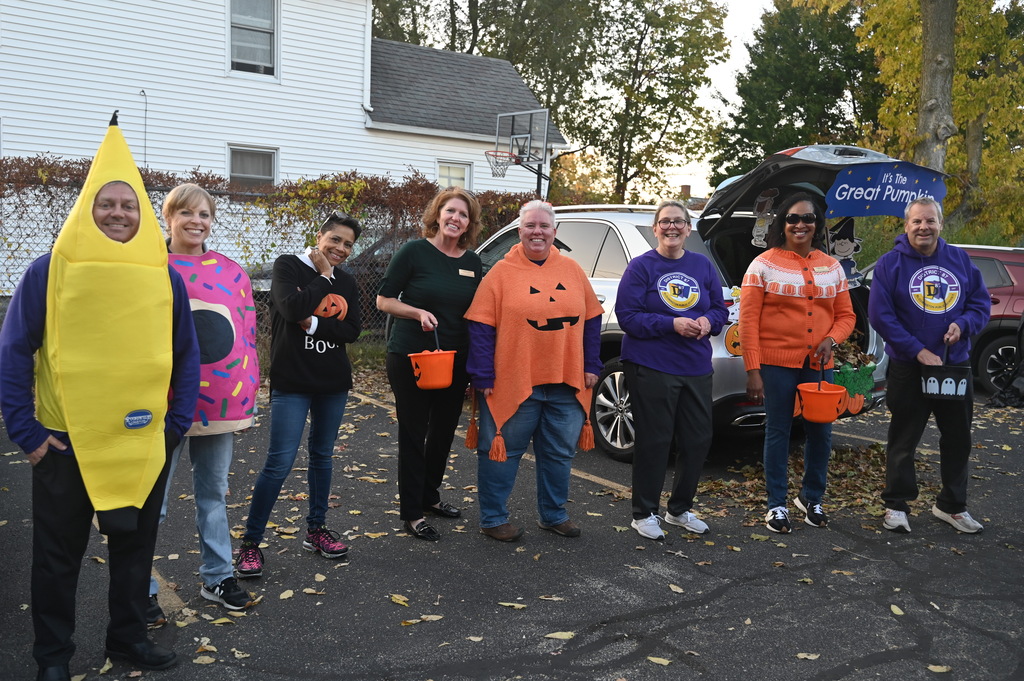 Group of adults in Halloween costumes standing next to a vehicle. Leaves on the ground.