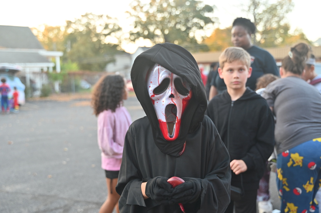 A child in a mask walks with others in a parking lot. They hold a red object.