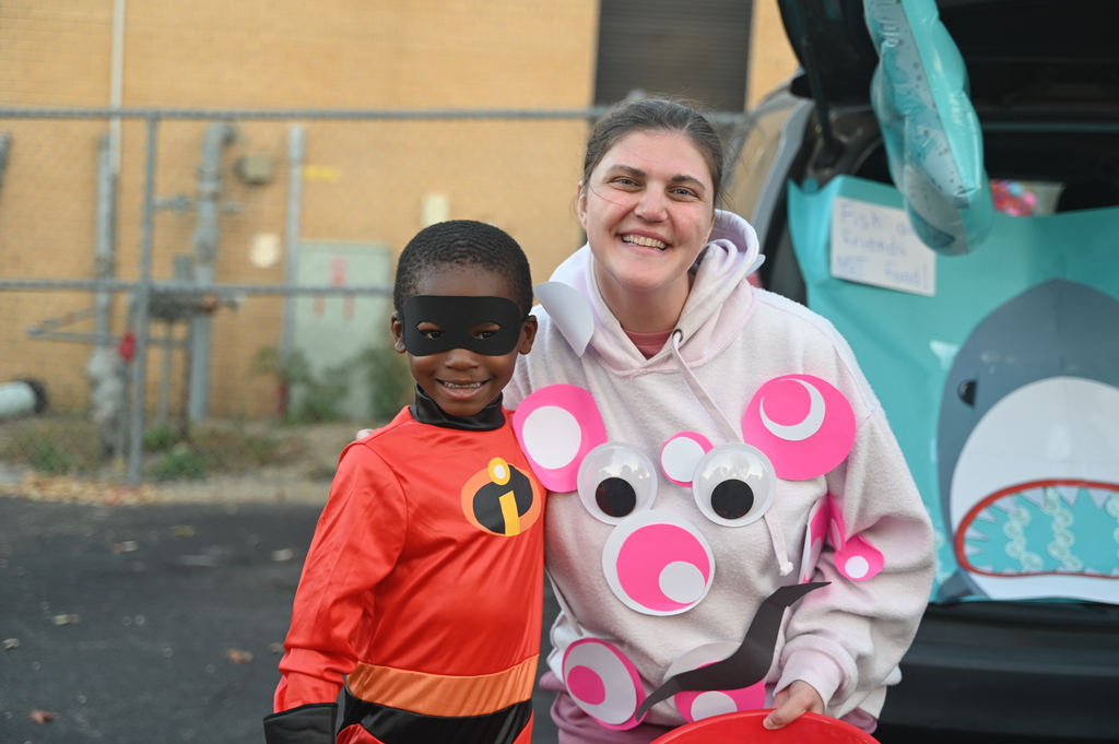 Two people stand by a car; a boy in a red costume and a woman in pink with pig ears.