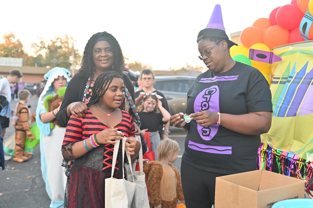 A woman and a girl in costumes stand near a decorated booth with a person in a crayon costume.