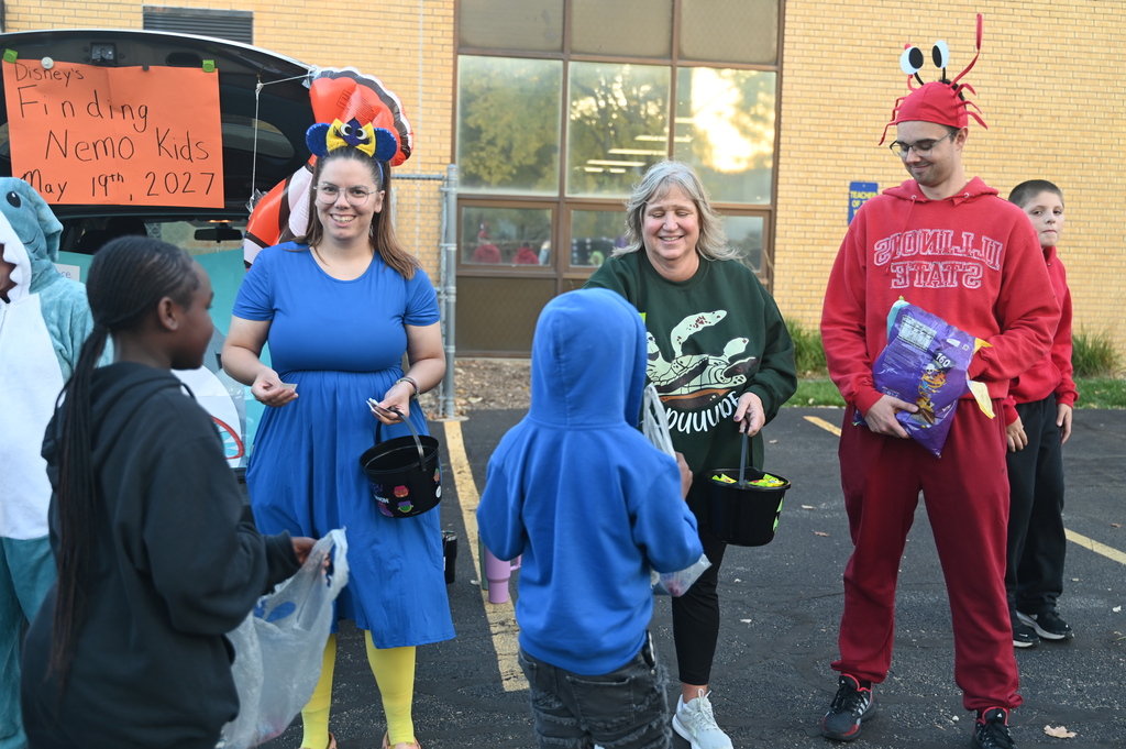 Several people, some in costumes, stand outside near a car with a banner. They hold bags and buckets.