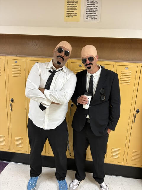 Two people wearing costumes and sunglasses stand in front of yellow lockers in a school hallway.