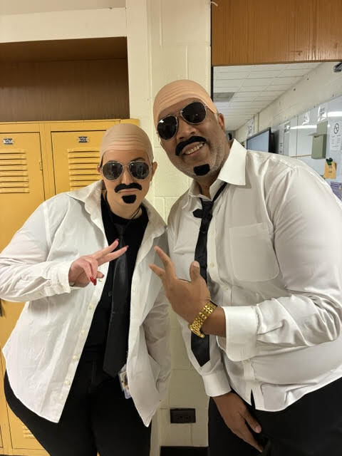 Two men wearing white lab coats and sunglasses with mustaches pose in front of lockers.
