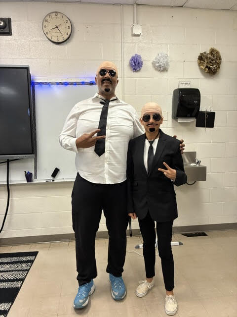 Two people in suits and sunglasses stand close together. One is taller, the other shorter. Behind them is a whiteboard and a sink.
