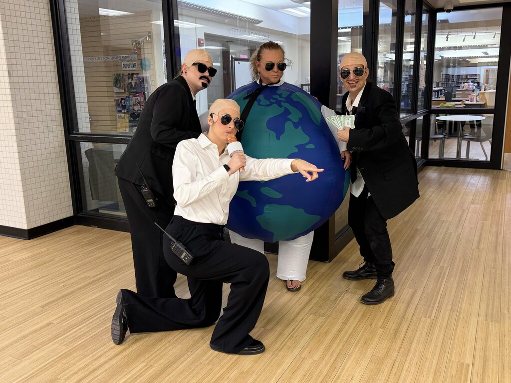 Four people in suits and sunglasses pose in a room; one crouches, one holds a globe.