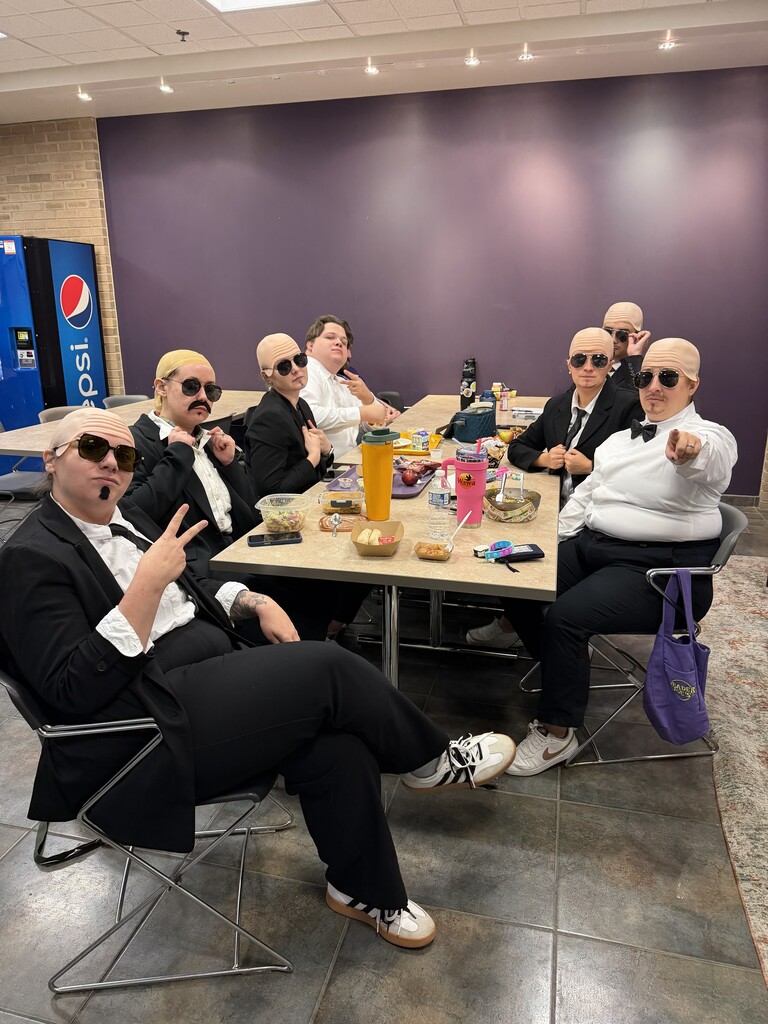 Nine people in suits and sunglasses sit around a table with food and drink in a cafeteria.