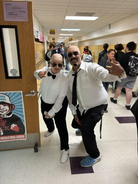 Two men dressed in white shirts, bow ties, and sunglasses stand in a hallway, posing for a photo.