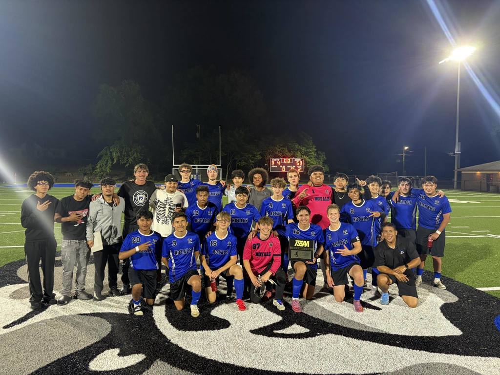 A high school boys soccer team from Eagleton College and Career Academy posing together on a turf field at night under bright stadium lights. The players are wearing blue "Royals" jerseys and black shorts, with two goalkeepers in bright pink jerseys. One player in the front row is holding up a TSSAA District Champion trophy.