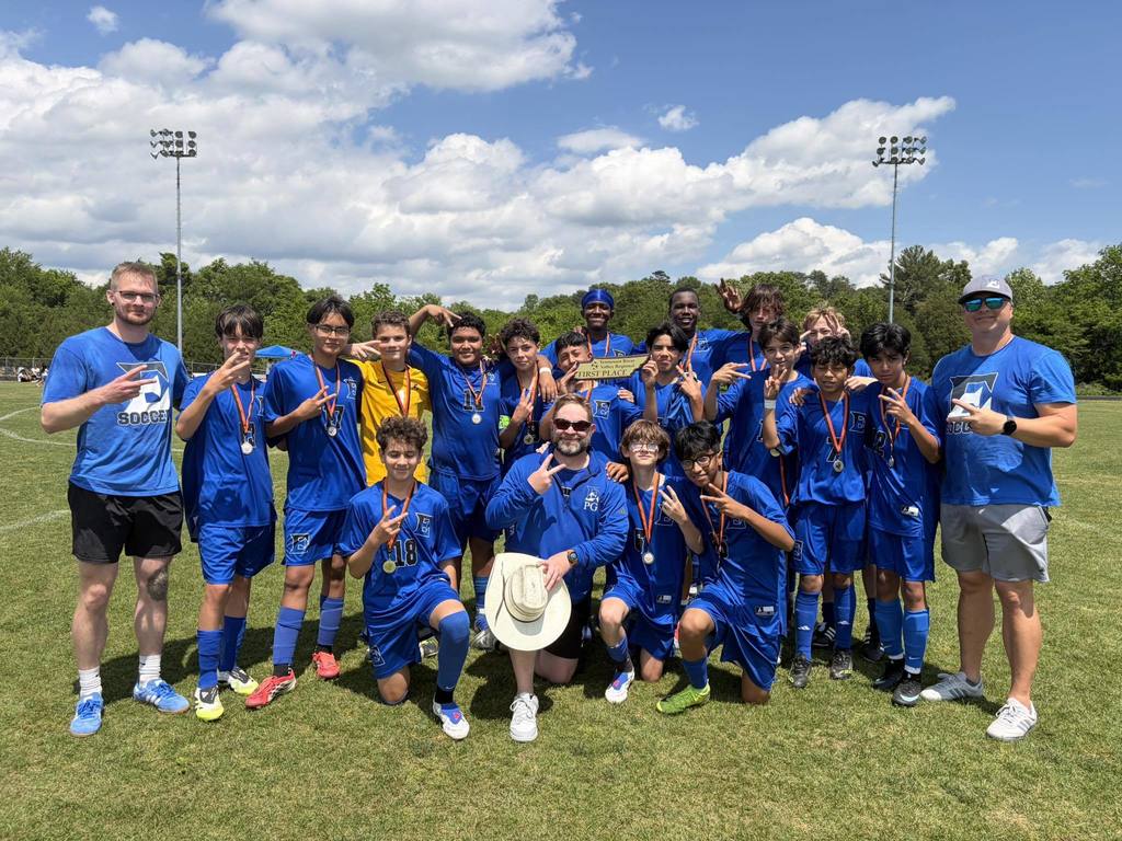 A middle school boys soccer team and their coaches from Eagleton College and Career Academy posing on a sunny grass field. The players are wearing blue jerseys with an "E" logo and many are wearing winner's medals around their necks. Two players in the center hold a wooden "First Place" trophy.