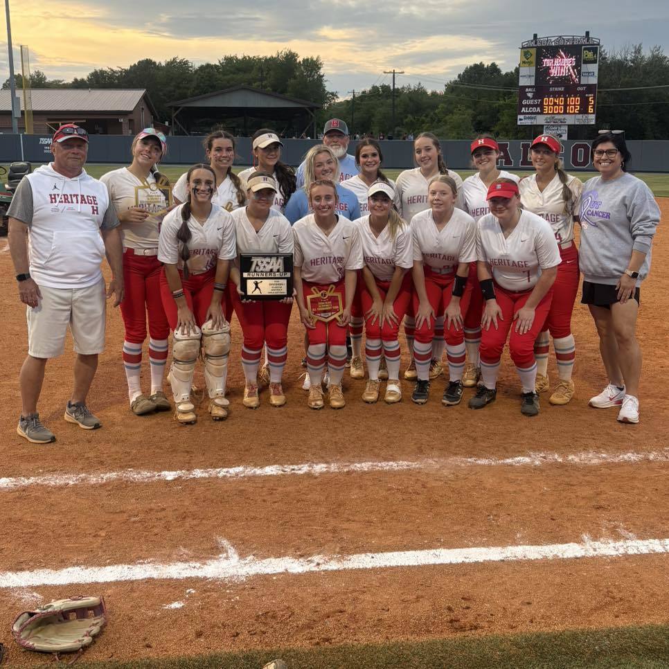 A high school girls softball team and their coaches from Heritage High School standing on a red clay infield. The players are wearing white jerseys with "Heritage" in red and red pants. Two players in the center hold a TSSAA Runners-Up plaque and a district tournament award.