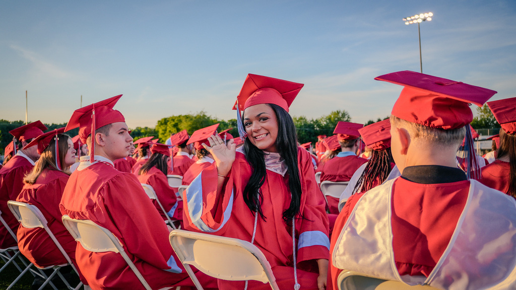 candid, close-up photo of a female graduate with long dark hair wearing a red cap and gown, smiling and waving at the camera while seated among her peers on a football field.