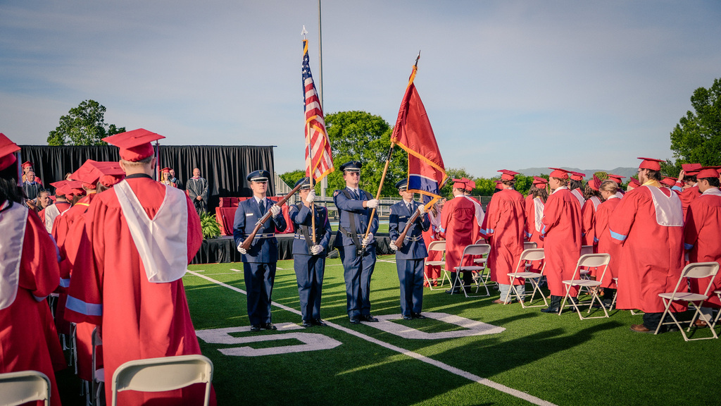 An Honor Guard composed of four individuals in blue uniforms standing at the 50-yard line of a football field, holding the American flag and a school flag during the graduation ceremony.
