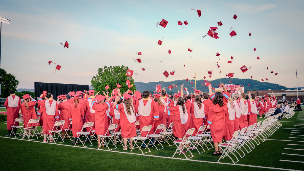 A wide, low-angle shot of a large group of graduates in red gowns and caps standing on a green athletic field, joyfully tossing their graduation caps into the air at dusk with mountains in the background.