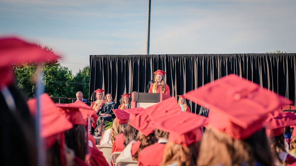 A view from behind the seated graduates, focusing on a student speaker at a podium on a stage with a black curtain backdrop, framed by the blurred red graduation caps of students in the foreground.