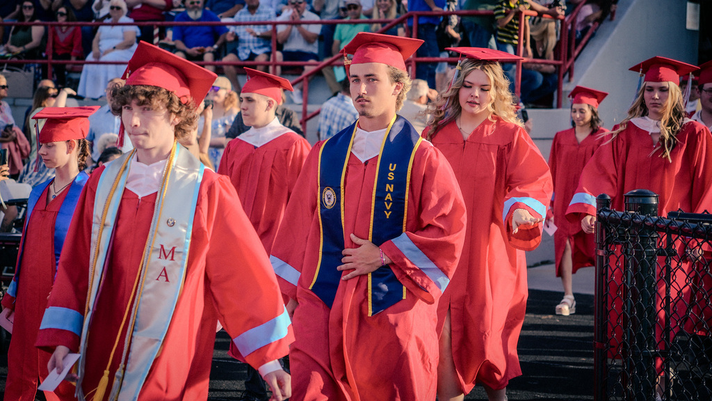 A group of graduates processing onto the field; in the center, a male student wears a red gown with a navy blue "US Navy" stole, followed by a female student in a red gown.