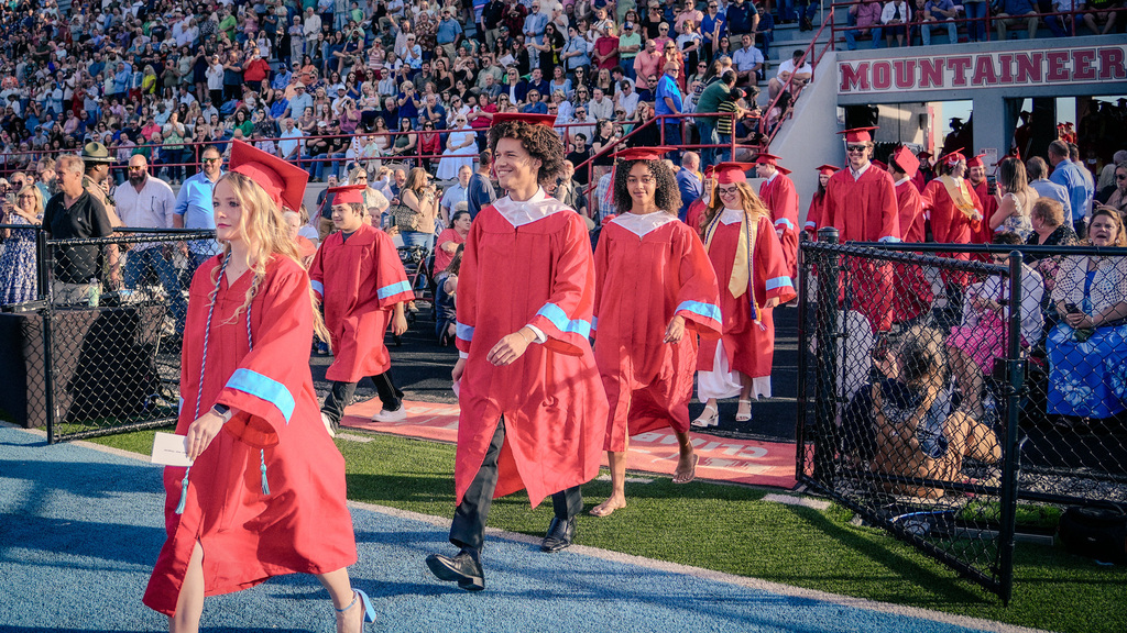 Graduates in red gowns and caps walking onto a green athletic field through a gate, passing a "Mountaineers" sign on the stadium wall while spectators cheer from the bleachers.