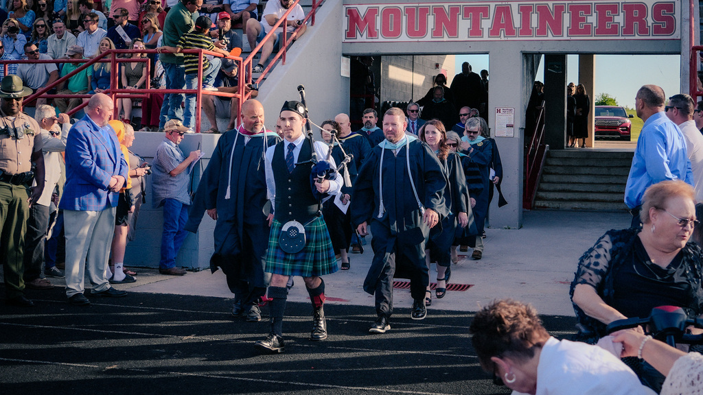 The start of the graduation procession featuring a bagpiper in a traditional green kilt and vest leading faculty and administrators in dark academic regalia onto the athletic field.