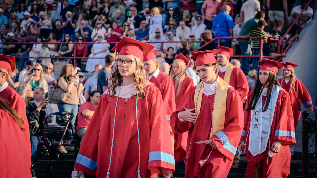 A close-up of graduates in a procession; a female student with glasses and a red gown leads the group, followed by a male student and a female student wearing a light blue "CNA" stole.