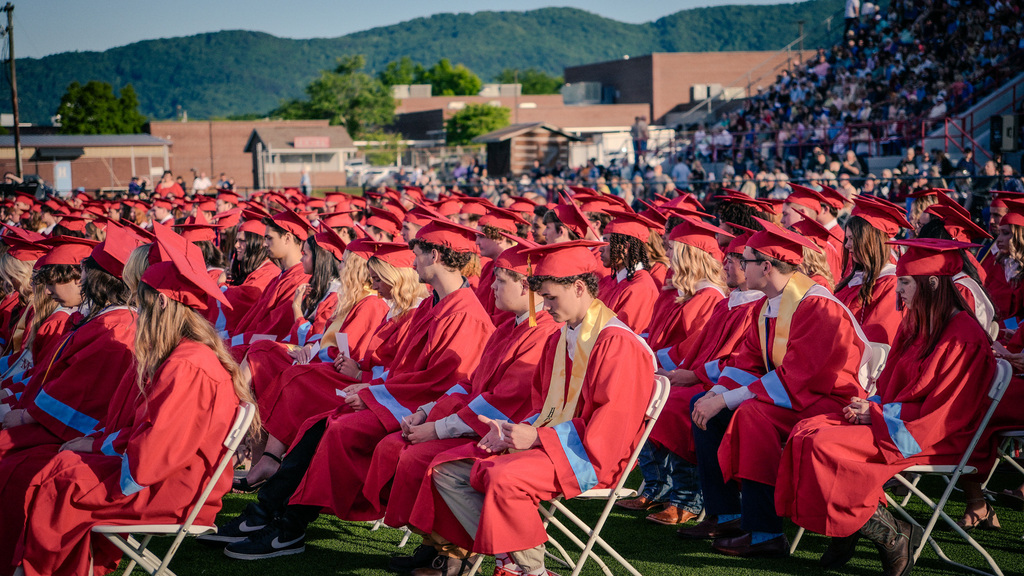 A side view of several rows of graduates in red gowns and caps seated in white folding chairs on a green field, looking toward the stage with a large crowd in the bleachers and blue mountains in the distance.