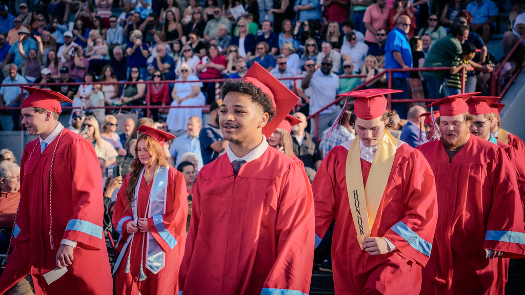 Graduates walking in a procession past the bleachers; one student in the foreground wears a red gown and a gold "Honor" stole, while a large crowd watches from the stands behind them.