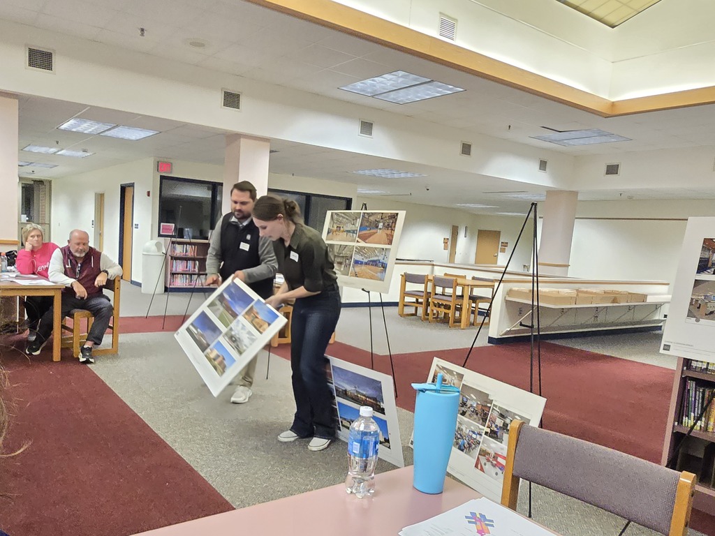 Interior of a library with tables and chairs. People seated at a table, and others standing in the center.