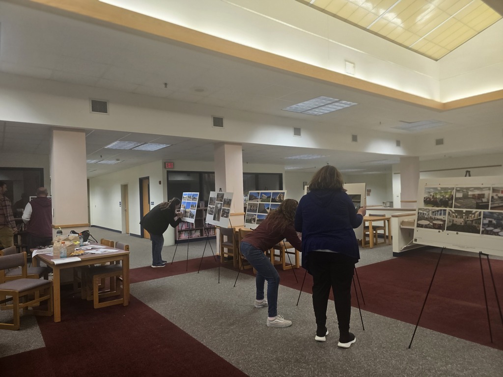 Two adults examining artwork on easels in a room with tables, chairs, and a red carpet.