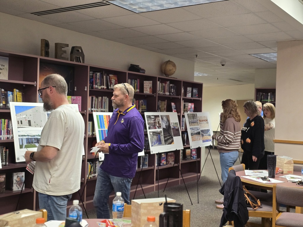 Adults stand around room with bookshelves, posters on easels, and table with jacket on it.