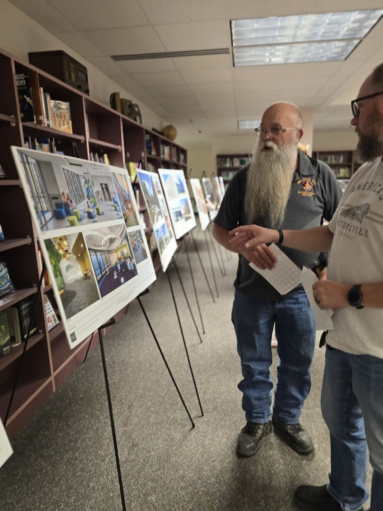 Two men examine architectural drawings displayed on stands in a library, surrounded by tall shelves filled with books.