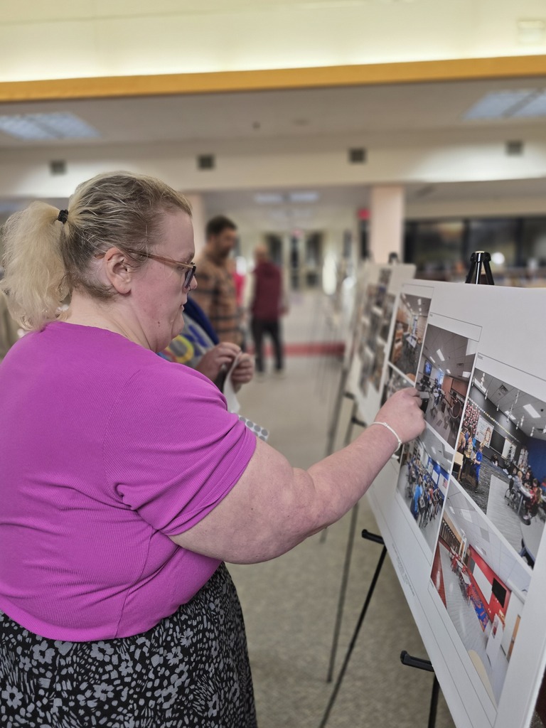 A woman in a purple shirt and glasses looks at photos on a board in a school.