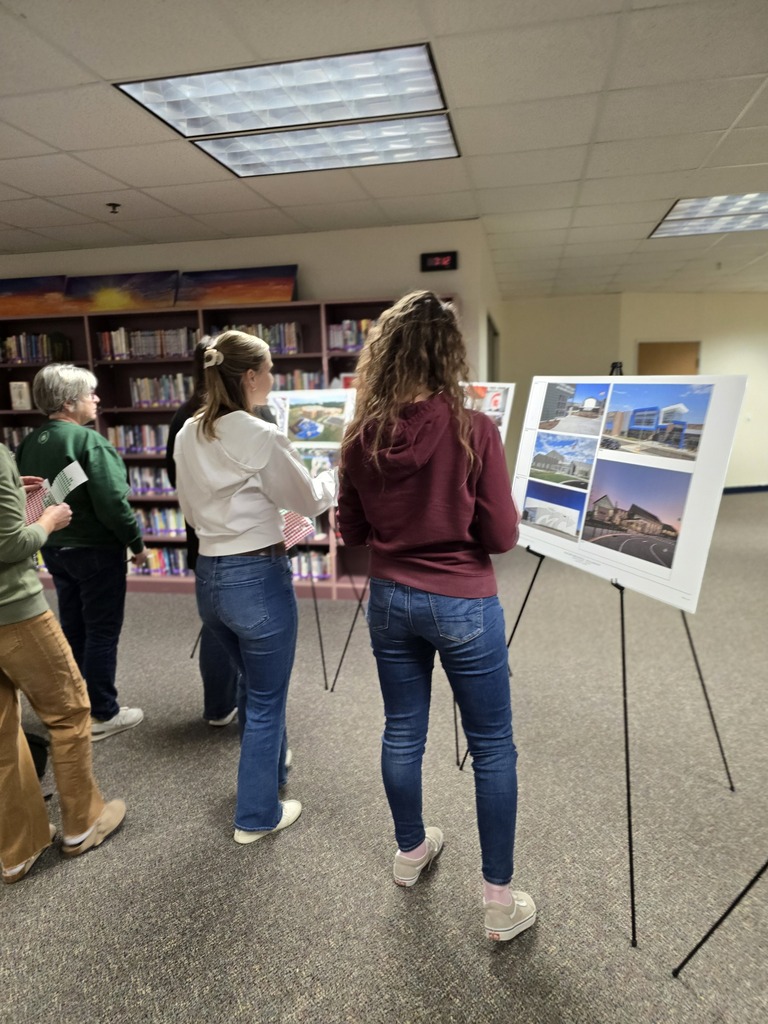 Five people stand in a room, examining architectural drawings on display stands. Shelves filled with books are in the background.