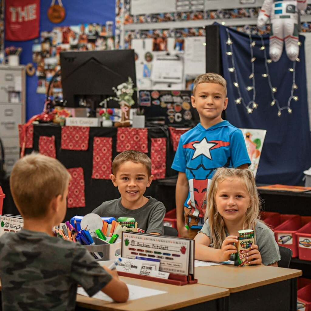 Four children in a classroom with a decorated background. Three seated, one standing, holding a can.