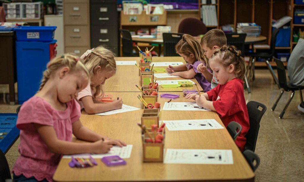 Children in a classroom, seated at a table, working on paper with markers and crayons.